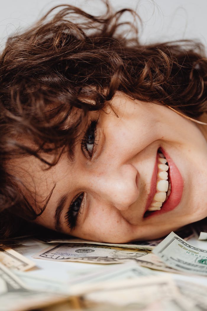 Caucasian woman with curly hair smiling while lying on a pile of paper money.